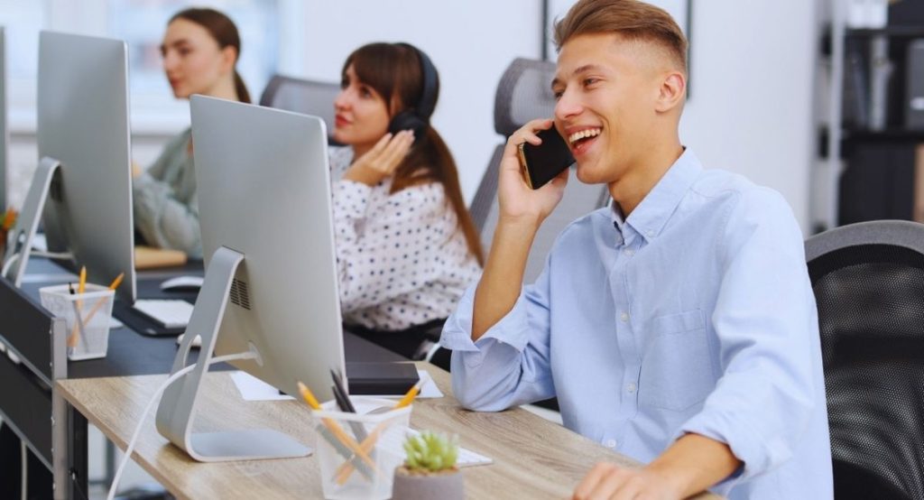 Jeune commercial souriant au téléphone devant son ordinateur, collègues en appel en arrière-plan
